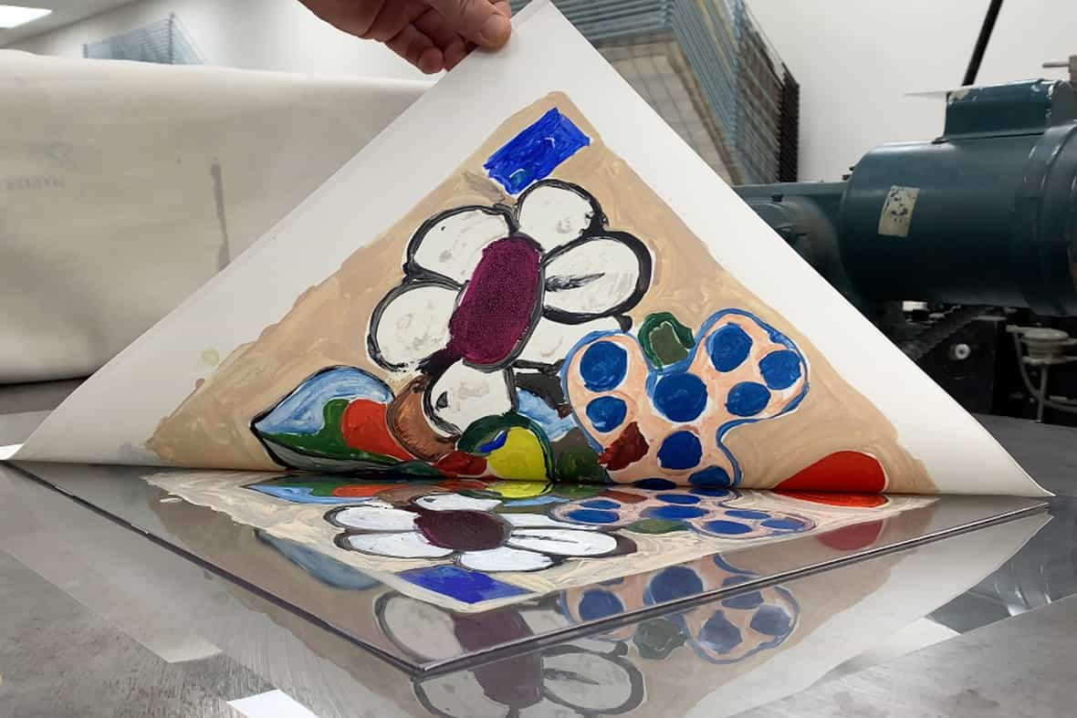 Close up image of a man pulling up the corner of a sheet of paper, revealing a printed image made using an etching press.