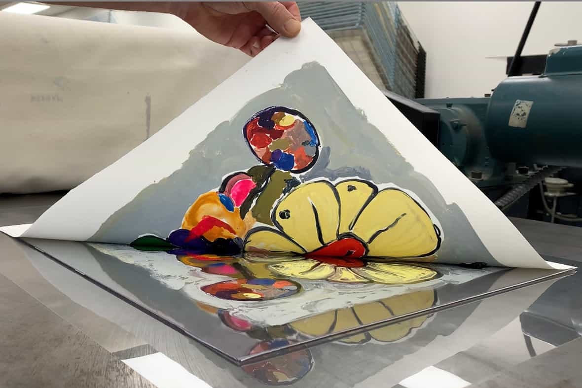 Close up image of a man pulling up the corner of a sheet of paper, revealing a printed image made using an etching press.