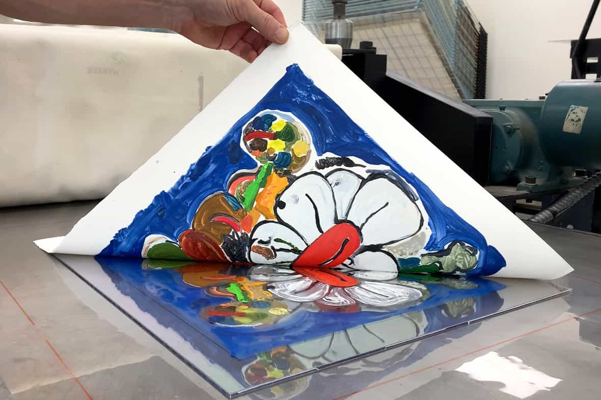 Close up image of a man pulling up the corner of a sheet of paper, revealing a printed image made using an etching press.